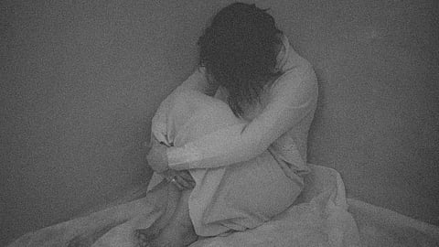 An image of a girl sitting at the corner of bed and hugging her knees and hising her face.