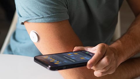 Image of a man in teal shirt using smartphone to check blood glucose via a healthcare wearable device on his arm.