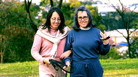 A young woman walks arm-in-arm in a park,with an older woman smiling and chatting.They wear comfortable exercise clothing, surrounded by trees and green lawns.