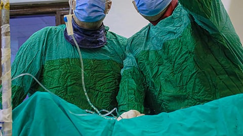 Two surgeons in green scrubs and masks, with one pointing to a monitor. IV lines hang above.