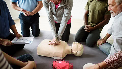 A group of people attentively watch a woman performing CPR on a mannequin. A red first aid kit lies nearby.