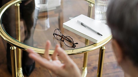 A book placed on a table with a pen and spectacles beside it, while a man conducts a consultation in an office setting.