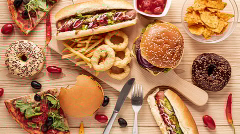 Image of fast food on a wooden table, including burgers, hot dogs, pizza, onion rings, fries, donuts, and nachos.