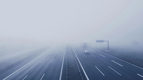 Dense fog and heavy pollution shroud a busy road with cars and a traffic light.