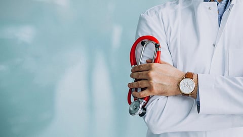 A doctor in a white coat holds a red stethoscope, arms crossed.