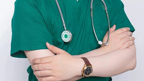 Close-up of a doctor wearing dark green scrubs with a stethoscope around their neck and a brown wristwatch visible.
