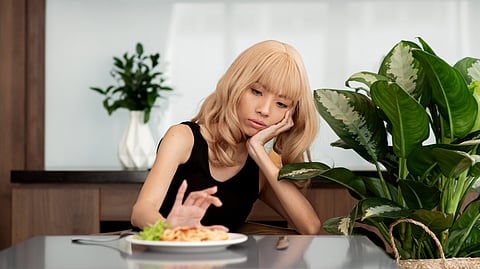 A person with blond hair in a black tank top looks thoughtfully at a plate of food.