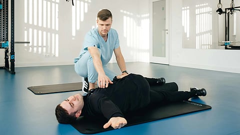 Healthcare physician in blue scrubs assisting a male patient with guided physical rehabilitation exercises in a clinical setting.