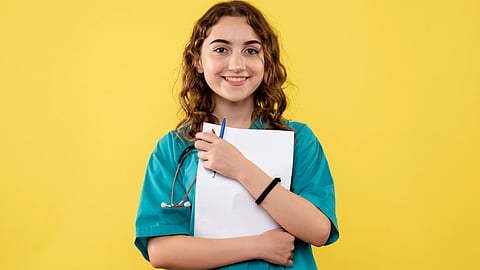 Medical student in blue uniform, stethoscope around neck and paper and pen on hand.