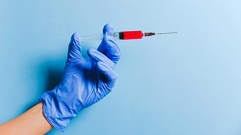 A hand in a blue medical glove holds a syringe with red liquid.