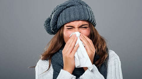 Woman in winter clothing sneezes into a tissue. She wears a gray knit hat and sweater.