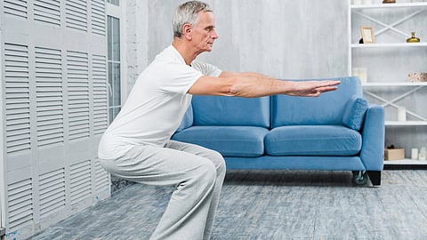 An older man exercise in a bright room, performing a squat with arms extended.