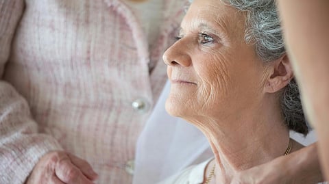An older woman with gray hair looks thoughtfully into the distance while another person gently supports her from behind.