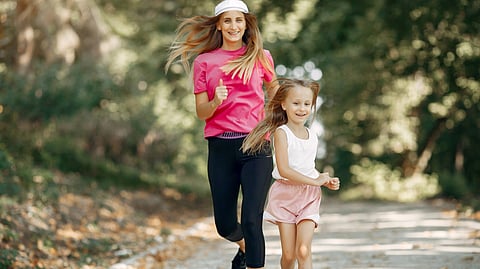 A woman in a pink shirt and a girl in a white top joyfully jog on a sunlit forest path.