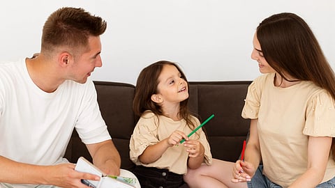 A family of three sits on a couch, happily engaged, child smiles between the parents.