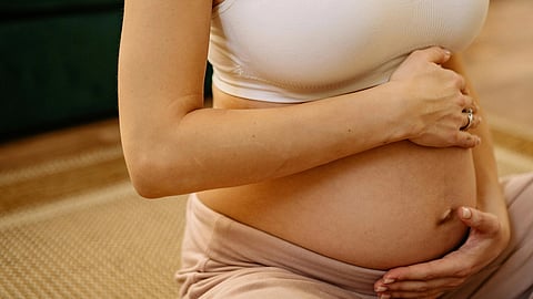 A pregnant woman gently holding her belly, wearing a white crop top and beige trousers.