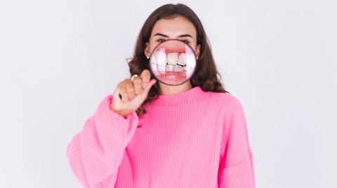 Young woman in pink sweater on white wall background with magnifier shows white teeth zoomed in. 