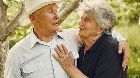 An elderly couple standing in a forest, looking at each other; the woman wears a navy blue t-shirt, and the man wears a white shirt.