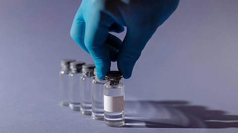 A gloved hand picks up a vial from a row of clear liquid vials on a light purple surface.