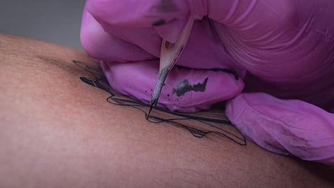 Close-up of a tattoo artist's gloved hand, wearing pink gloves, as a needle outlines a black design on skin. 