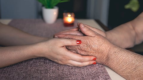 Two hands gently hold an elderly hand over a table with a textured cloth.