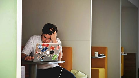 A visibly stressed man sitting alone in a café, hunched over his open laptop with his head in his hands.