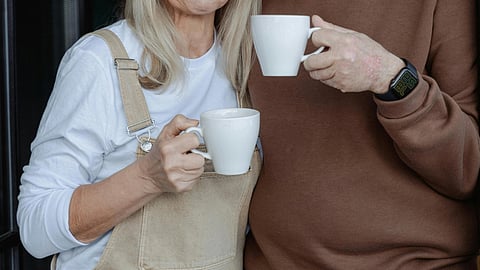 Elderly couple enjoying coffee together, the man wearing a brown hoodie and the woman in a beige dress.
