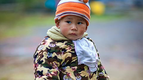 A child wearing a jacket and a cap, standing outdoors against a blurred background.