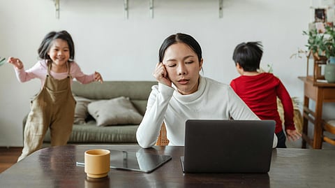 Women looking stressed while working on laptop and two kids playing in the background.
