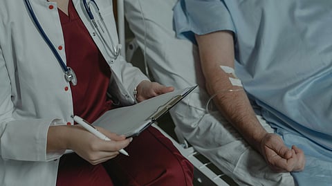 A nurse in maroon scrubs, apron, and stethoscope takes notes on a patient’s medical history at the bedside.