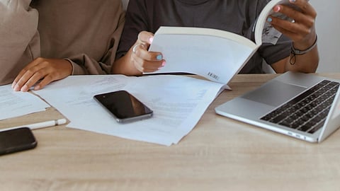 Two women at a table reviewing documents, with a laptop and a phone on the table, and one woman holding a book.