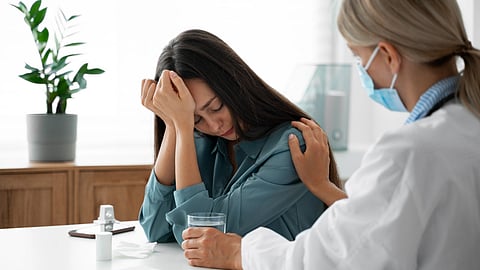 A woman looks distressed, holding a glass of water, while a doctor attends to her.