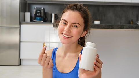 Smiling woman in a blue top holds a white supplement bottle and a capsule.