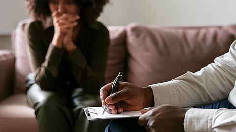 A person sits on a sofa with hands clasped while another takes notes on a clipboard.