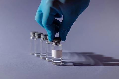 Close-up of a man holding a vaccine vial with a blank white label, with several other vaccine vials lined up behind it against a purple background.