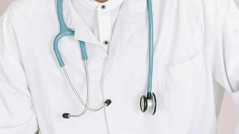 Close-up of a blue stethoscope with a doctor wearing a white shirt and apron in the background.