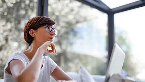 A woman wearing a white top and blue spectacles sits in front of her laptop, resting her chin on her hand as she appears deep in thought.