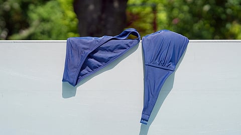 Blue bikini top hangs over a white fence against a blurred leafy background.