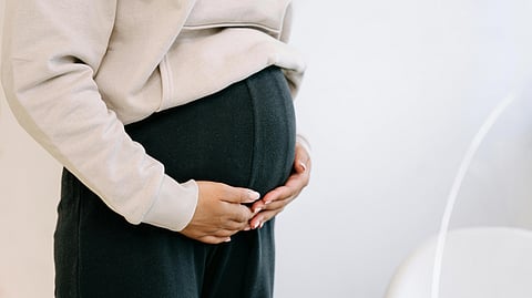 A pregnant woman wearing black leggings and a beige hoodie, gently holding her belly