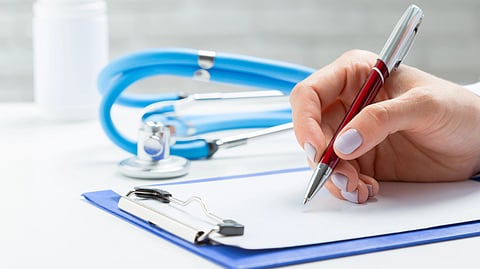Hand with purple nails writing on a clipboard, with stethoscope and bottle behind.