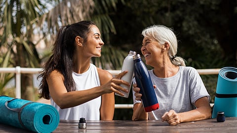 Two women, one older and one younger, smile and clink water bottles at an outdoor table. 