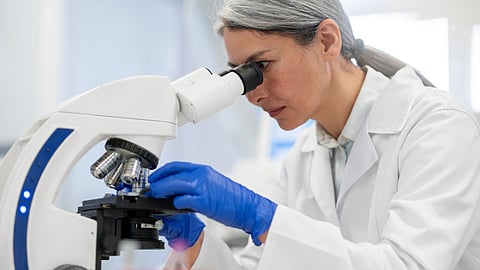A scientist in a white lab coat and blue gloves peers into a microscope.