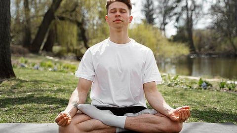 A person in a white T-shirt meditates in a cross-legged pose on a mat by a lake.