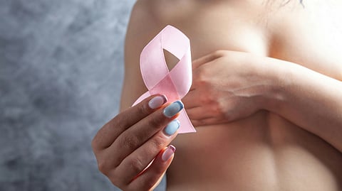 A woman with pastel nail polish holds a pink breast cancer awareness ribbon against her chest.