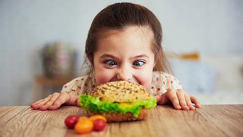 A young girl with an excited expression stares eagerly at a large sandwich with lettuce on a wooden table.