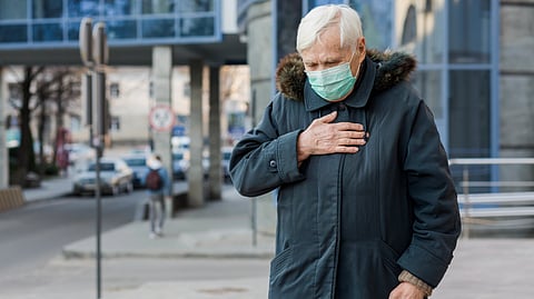 Elderly man wearing a green face mask and dark coat holds his chest.