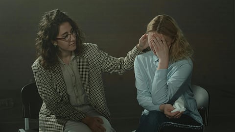 Two women seated together, one in a light blue top holding her head with a sad expression while another woman wearing glasses sits beside her, offering comfort and support.