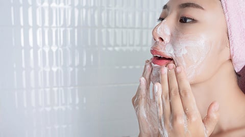 A person in a pink towel headband is applying a foamy facial cleanser in a tiled bathroom.