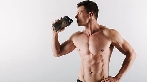 Muscular man drinking from a sports bottle, standing shirtless.