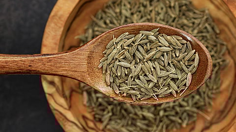 A close-up of a wooden spoon filled with cumin seeds resting over a wooden bowl.